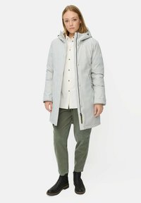 Lichtgrijze gewatteerde jas met een capuchon, ritsvoering, drukknoopsluitingen en zijzakken. Draagt over een wit shirt en olijfgroene broek, met zwarte enkellaarsjes.