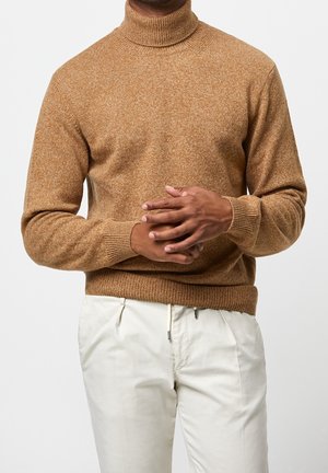 Mand iført en brun rullekravesweater og off-white bukser med snøre, med hænderne foldet foran livet.