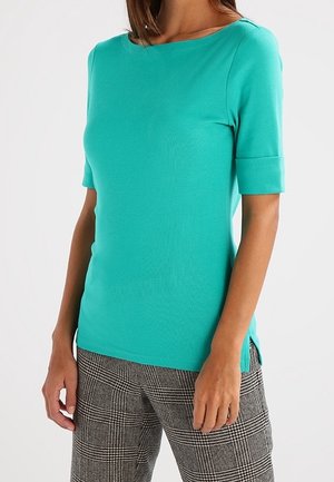 Kobieta w turkusowej bluzce z krótkim rękawem i czarno-białych spodniach w kratę, widoczna od szyi do bioder na białym tle.