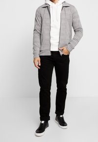 Giacca grigia con zip e motivo a quadri sopra un felpa bianca con cappuccio, abbinata a jeans neri e sneakers nere con suole bianche.