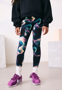 Leggings sötét alapon, többszínű absztrakt csavarodó mintával. Lila velúr sportcipővel és fehér texturált zoknival párosítva.