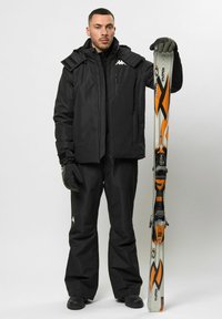 Schwarze wasserdichte Skijacke mit hohem Kragen und verstellbarer Kapuze, kombiniert mit passenden schwarzen Hosen. Orange- und graue Skier in der linken Hand gehalten.