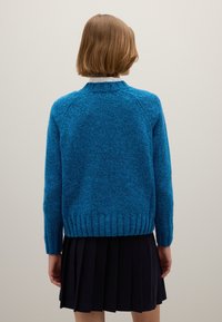 Blauwe gebreide trui met een gestructureerde afwerking en geribbelde zoom, voorzien van een ronde halslijn en lange mouwen. Draagt over een geplooide zwarte rok.