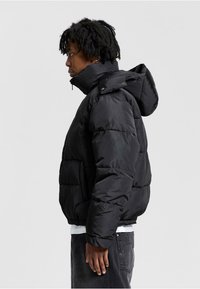 Sort pufferjakke med høj krave og justerbar hætte. Har kraftig polstring, lynlåslukning foran og sidelommer.