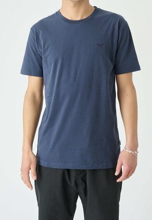 Mężczyzna noszący jednolity granatowy t-shirt z krótkim rękawem i czarne spodnie, z srebrną bransoletką i pierścionkiem na prawej ręce.