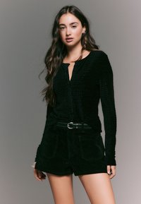 Top nero a maniche lunghe con trama, apertura a goccia sul davanti e chiusura con bottone, abbinato a pantaloncini neri in velluto a coste a vita alta e cintura nera.