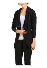 Zwarte gebreide cardigan met een open voorkant, geribbelde manchetten en een zachte textuur. Gedragen over een wit shirt en gecombineerd met donkere broek.