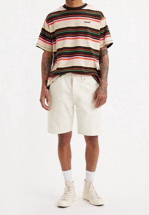 T-shirt Levi's à manches courtes rayé en crème, rouge, vert et noir ; associé à un short en jean blanc et des baskets montantes beiges avec des chaussettes blanches.
