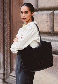 Schwarze Wildleder-Tote mit glatten Lederelementen, mit einem abgerundeten oberen Teil und geprägt Logo an der Unterseite. Das Model trägt ein helles Hemd.
