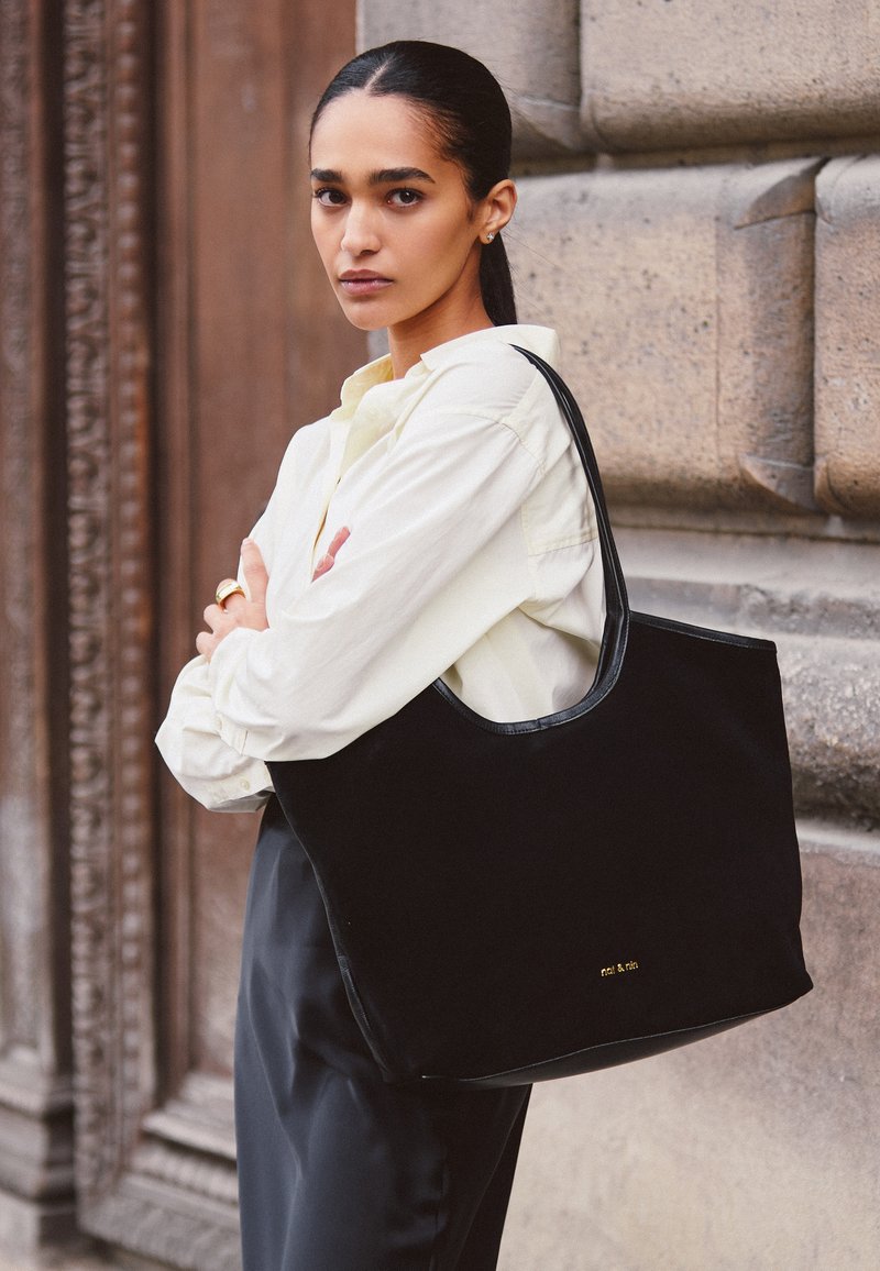 Schwarze Wildleder-Tote mit glatten Lederelementen, mit einem abgerundeten oberen Teil und geprägt Logo an der Unterseite. Das Model trägt ein helles Hemd.