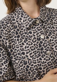 Camicia maculata in tessuto morbido, con colletto e bottoni a pressione argentati. Macchie nere su uno sfondo beige con finitura testurizzata.