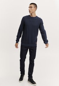 Alva tamsiai mėlyna megztinis su apvaliu iškirpimu, raukiniais rankogaliais ir apačia. Derinamas su tamsiais džinsais ir juodais sportbačiais.