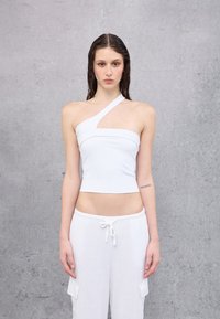 Top blanco de un solo hombro, corto y fabricado en un tejido suave; presenta un corte diagonal y un diseño ajustado. Combinado con pantalones blancos sueltos de cordón.