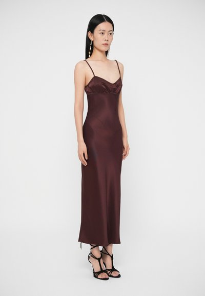 Žena v bezrukávových hlbokých bordových saténových šatách so štíhlymi ramienkami a čiernych sandáloch na vysokom opätku so remienkami, stojaca pred bielym pozadím.