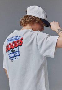 Jeune personne aux cheveux bouclés portant un t-shirt blanc avec le texte « Worker Goods Service » et une casquette blanche inclinée vers l'avant, montrant le design au dos.