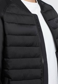 Giacca bomber nera imbottita con una texture liscia, caratterizzata da una chiusura con zip, colletto a costine e dettaglio del logo. Design semplice e lineare.