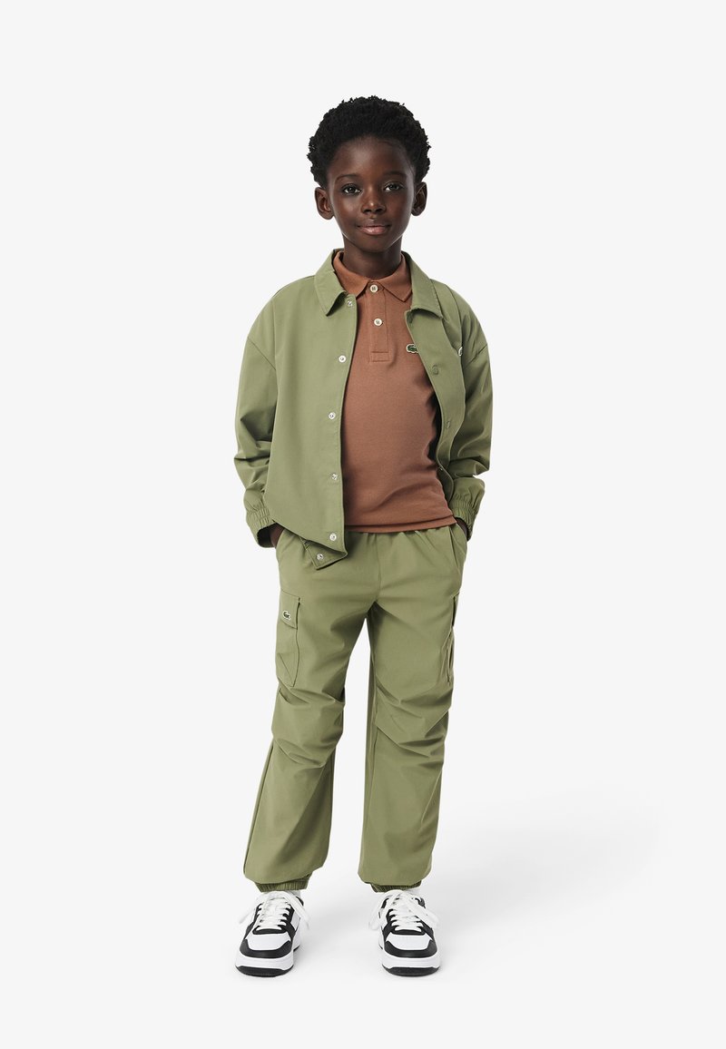 Giacca verde con bottoni a pressione, polo marrone e pantaloni verdi abbinati. Caratteristiche includono polsini elasticizzati e tasche laterali. Sneakers bianco e nero.