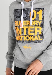 Sudadera gris con cordones blancos, que presenta un bloque de estampado de camuflaje amarillo y negro con el texto "01 Superdry International" en el pecho.
