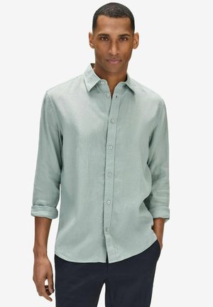 Mies, jolla on vaaleanvihreä pitkähihainen kauluspaita, hihansuut käännettyinä ja tummat housut, seisoo käsi yhdessä taskussa.