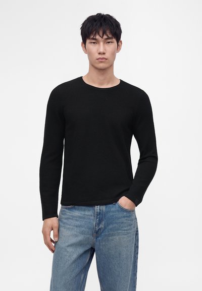 Jeune homme aux cheveux courts et noirs portant une chemise noire à manches longues et un jean bleu, debout avec une main dans la poche sur un fond blanc.