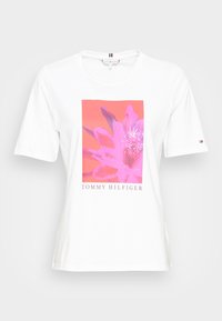 Camiseta blanca con un gráfico floral rosa y el texto "TOMMY HILFIGER" debajo. Mangas cortas, cuello redondo, corte casual.