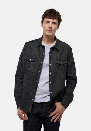 Chemise en denim noir avec deux poches poitrine et fermeture à boutons, portée par-dessus un t-shirt gris. Design ajusté avec des manches longues et une texture subtile.