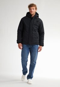 Giacca nera isolata con cappuccio, dotata di tasche con zip e una toppa con logo, abbinata a jeans blu e sneakers bianche.