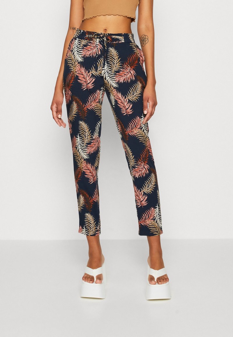 Femme portant un pantalon bleu marine avec des motifs de feuilles de palmier roses et beiges et des sandales compensées blanches, debout devant un fond uni.