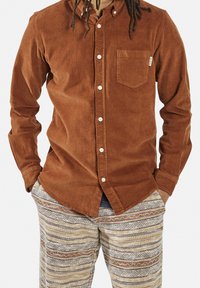 Camisa de pana marrón con cierre de botones y bolsillo parche, combinada con pantalones a rayas beige y marrón con estampado.