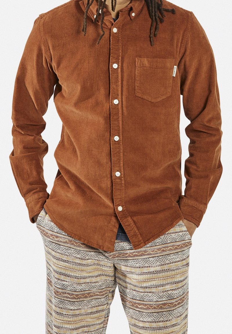 Camisa de pana marrón con cierre de botones y bolsillo parche, combinada con pantalones a rayas beige y marrón con estampado.