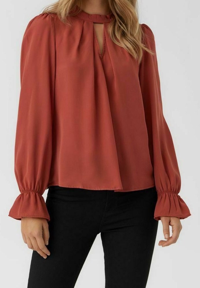 Blouse à manches longues de couleur rouille avec épaules bouffantes froncées, encolure en forme de goutte et poignets à volants portée avec un pantalon noir.