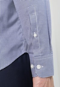 Koszula w niebiesko-białą kratę z długim rękawem, z mankietem zapinanym na dwa białe guziki, noszona na tle ciemnych spodni, pokazana na ramieniu osoby.