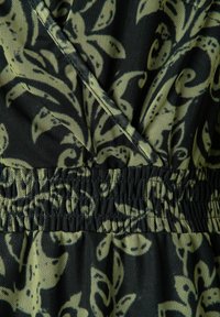 Zwarte stof met een gele bladachtige zwirls patroon, met een gathered elastische tailleband en een kruisontwerp aan de bovenkant. Textuurafwerking.