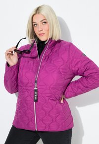 Žena s blond bob účesem, která nosí prošívanou fialovou bundu a drží černé sluneční brýle jednou rukou na bocích.