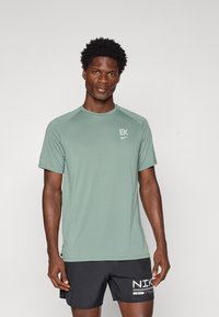 Ανοιχτό πράσινο αθλητικό μπλουζάκι με κοντά μανίκια, στρογγυλή λαιμόκοψη και λογότυπα σε διάφορα σημεία; συνδυασμένο με μαύρες βερμούδες με λεπτομέρειες λευκής επωνυμίας.