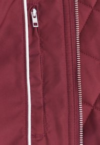 Bordeaux gewatteerde jas met een zilveren rits en een zijzak met witte afwerking. Glad textuur, minimalistisch ontwerp.