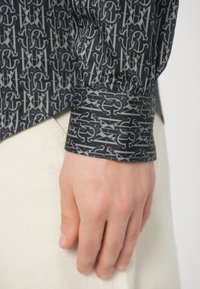 Koszula z długim rękawem wykonana z ciemnoszarej tkaniny z misternym, powtarzającym się wzorem logo. Mankiet rękawa prezentuje szczegółowy wzór.