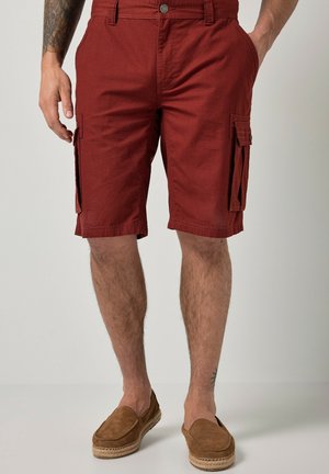 Mand iført rustfarvede cargoshorts og brune slip-on sko, står med hænderne i lommerne foran en ensfarvet baggrund.