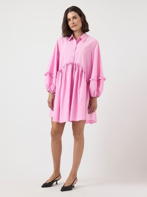 Woman in a loose, pink long-sleeve dress with ruffle details and black heeled slingback shoes, standing against a white background.