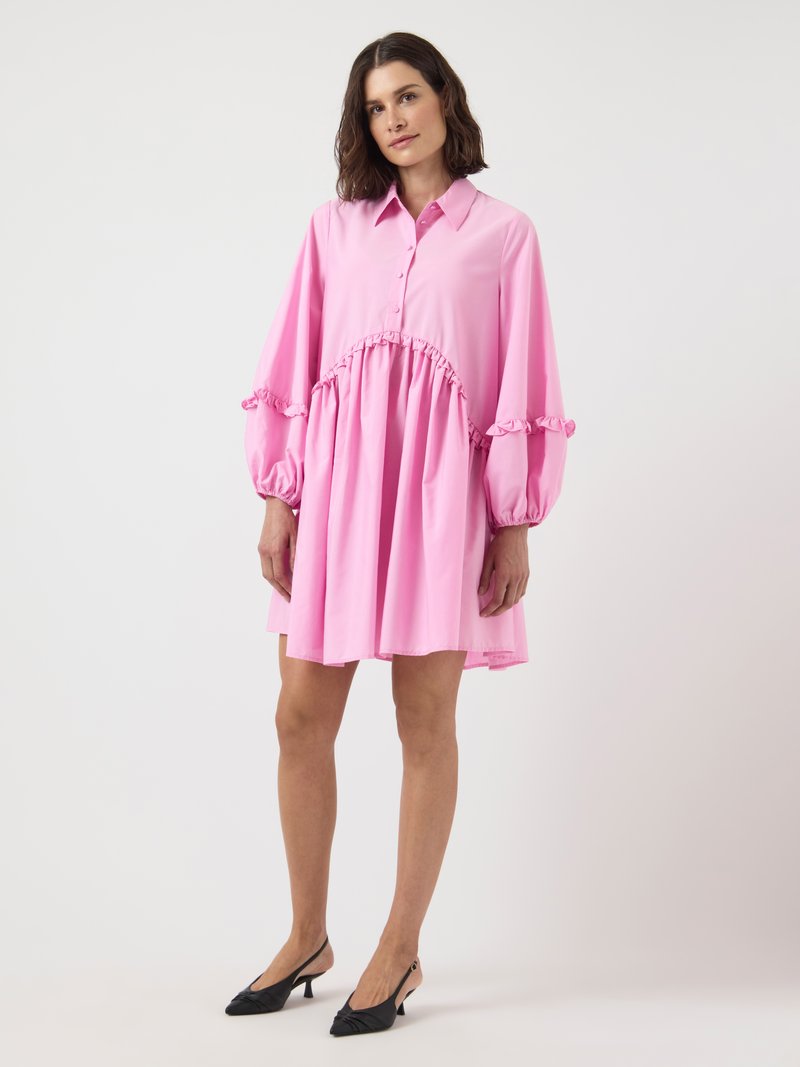 Kvinna i en lös, rosa långärmad klänning med volangdetaljer och svarta klackskor med slingback, stående mot en vit bakgrund.