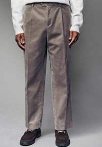Beige corduroy trousers with a straight leg design, featuring prominent vertical ribs and a zipper fly. Worn with a light sweater and dark shoes.
