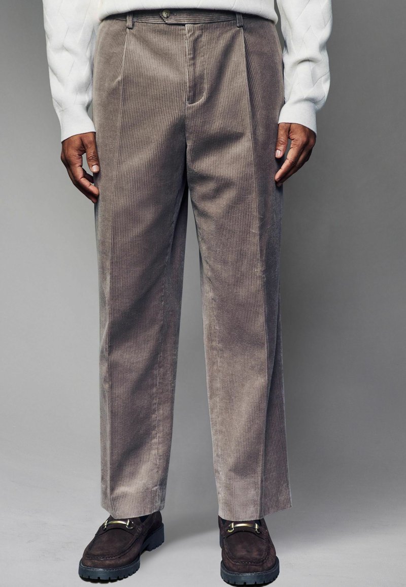 Beige corduroy trousers with a straight leg design, featuring prominent vertical ribs and a zipper fly. Worn with a light sweater and dark shoes.