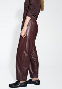 Pantaloni in pelle bordeaux con vestibilità comoda, cintura elastica e tasche laterali, abbinati a scarpe nere. Texture liscia e design sartoriale.