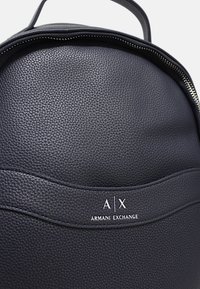 Zaino in pelle nera con texture, top arrotondato, dettaglio con zip e logo ricamato bianco "A|X" sul fronte.