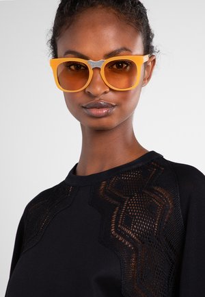 Donna con pelle scura e capelli raccolti che indossa grandi occhiali da sole arancioni e una maglietta nera con sezioni trasparenti e decorate vicino alle spalle.