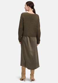 Frau mit dunklem Pferdeschwanz, die einen olivgrünen Strickpullover, einen passenden Midirock und beiges Wildleder-Stiefel bis zum Knie trägt, von hinten gewandt.