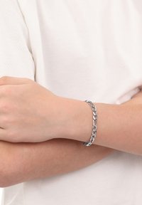 Bracciale in argento con maglie intrecciate, poggiato su un polso contro un indumento chiaro. Texture metallica liscia.