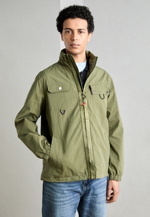 Green lightweight jacket with two chest pockets, elastic cuffs, and a front zipper. Features black and red hardware accents.