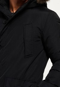Svart puffjacka med dragkedja och knäppning, avtagbar huva med fuskpäls, och en synlig ficka med patch. Mjuk, texturerad material.
