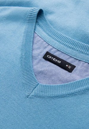 Lichtblauwe trui met een geribbelde V-hals. Beschikt over een label met de tekst "ombre" en een gladde textuur met een lichte glans.